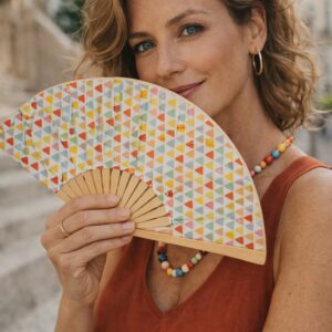Femme élégante tenant un éventail artisanal Sonia aux motifs géométriques colorés, dans une rue ensoleillée